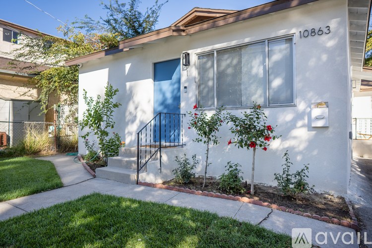 A white house with a blue door and windows is surrounded by greenery.