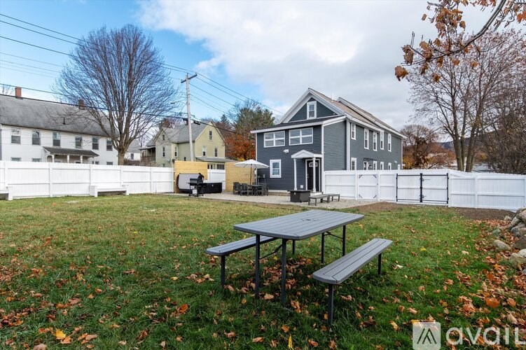 A picnic table is in the foreground of a grassy area with a white fence and a house in the background.