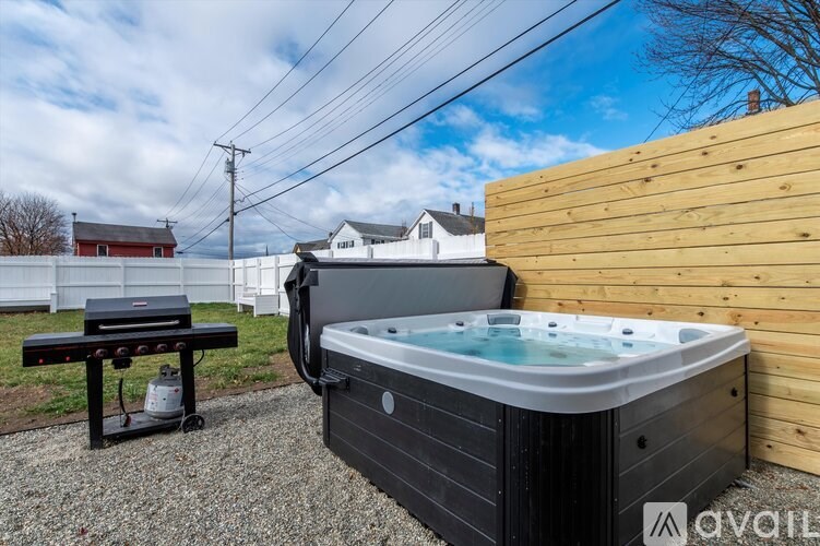 A hot tub sits on a gravel area next to a wooden fence and a grill.