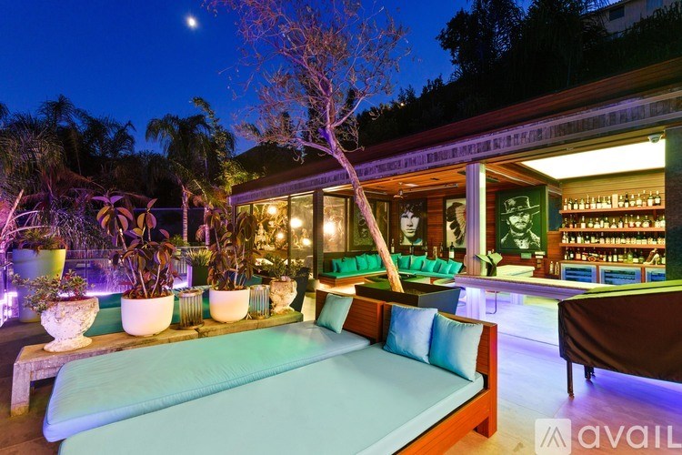 A cozy outdoor seating area with a long table and chairs, illuminated by warm lights, and a tree providing shade.