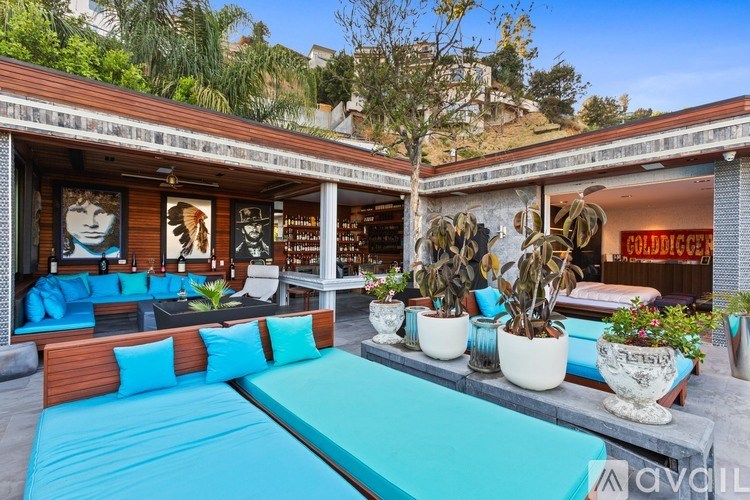 A pool table is surrounded by cushions and potted plants in a sunny outdoor room.