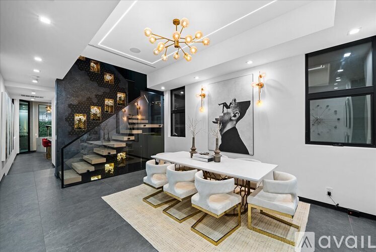 A modern dining room with a white table and chairs, a chandelier, and a staircase with a black and gold design.