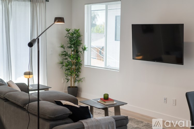 A living room with a grey couch, a black floor lamp, a small table with a green apple and a red book, and a flat-screen TV mounted on the wall.