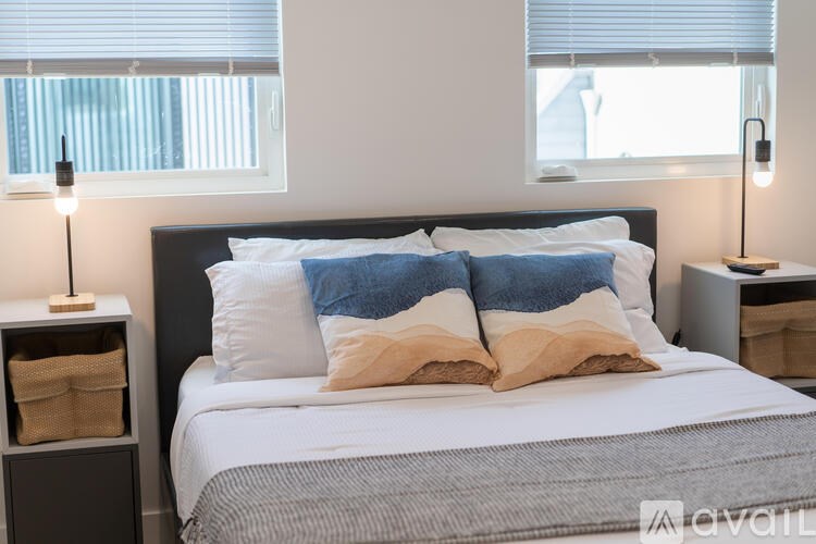 A bed with a white and grey comforter and two blue pillows is in the foreground with two lamps on bedside tables.