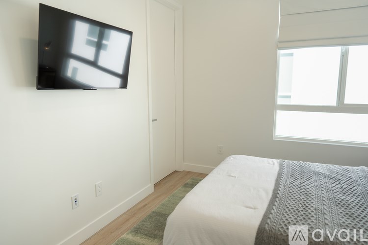 A bedroom with a bed, a flat screen TV mounted on the wall, and a window letting in natural light.