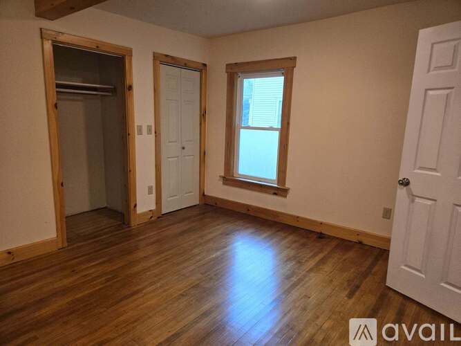 A room with wooden floors and white walls, with a door on the left and a window on the right.