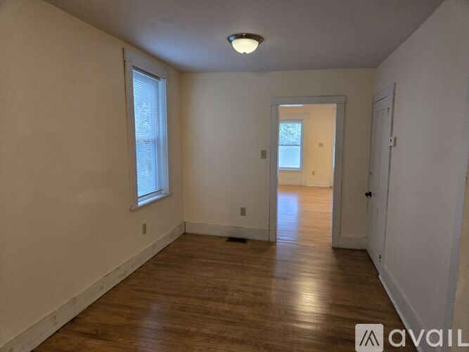 A room with wooden floors and white walls, with a window on the left and a door on the right.