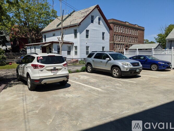 A white car is parked in a parking lot with other cars and a white house in the background.