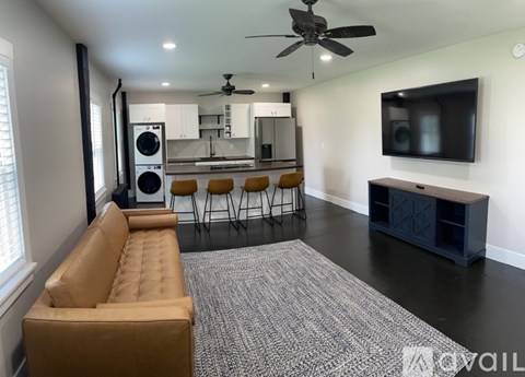 A living room with a brown sofa, a grey carpet, a ceiling fan, and a kitchen in the background.