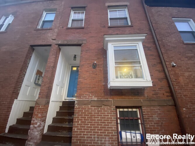 A red brick building with a white door and a window with a lit lamp.