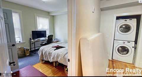 A bedroom with a bed, a desk with a computer, and a washing machine in the hallway.