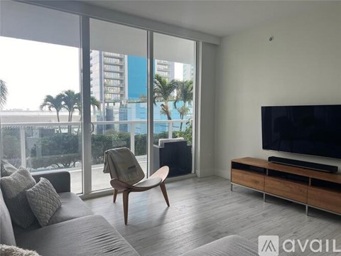A living room with a grey sofa, a wooden chair, a television and a large window overlooking a cityscape.