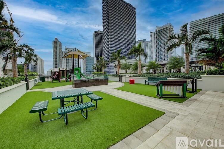 A park with green grass and picnic tables in the foreground with tall buildings in the background.