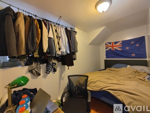 A bedroom with a bed, a desk with a laptop, a clothing rack with clothes, and a New Zealand flag on the wall.