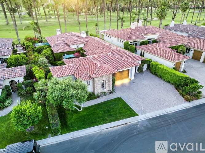A large house with a red tiled roof is surrounded by greenery and palm trees.