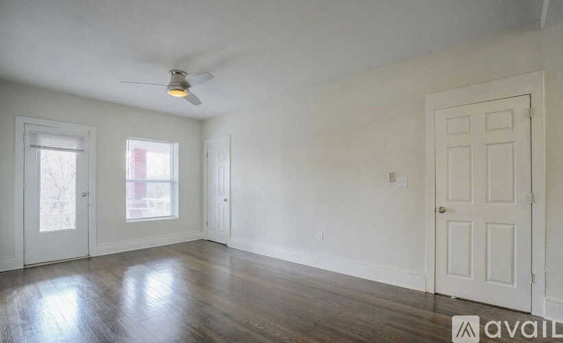 A room with a white door, a window with a view of a building outside, and a ceiling fan.