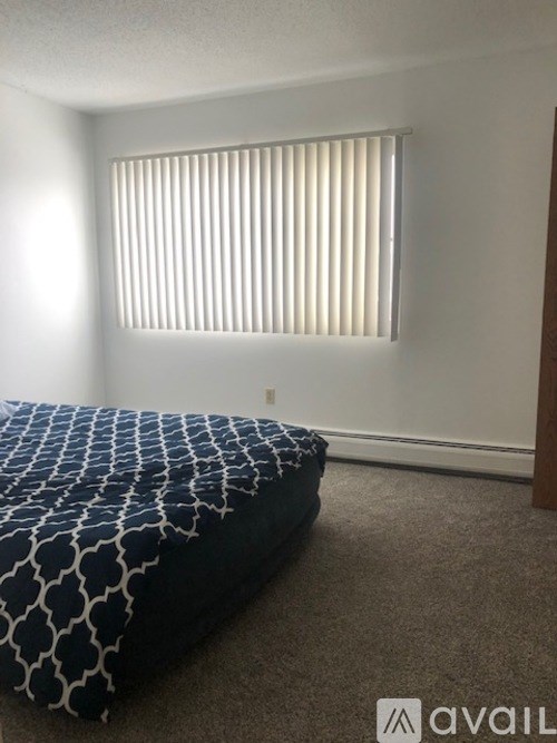 A bed with a blue and white patterned cover sits in a room with a window covered by blinds.