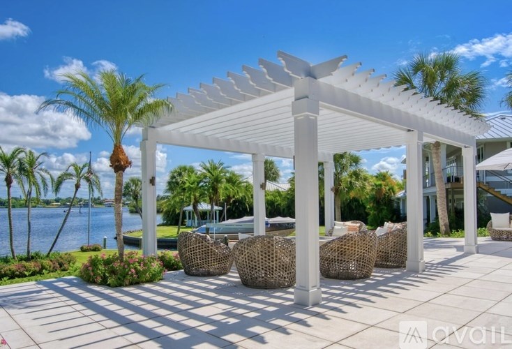 A white pergola with wicker chairs is set up on a patio with a body of water and palm trees in the background.
