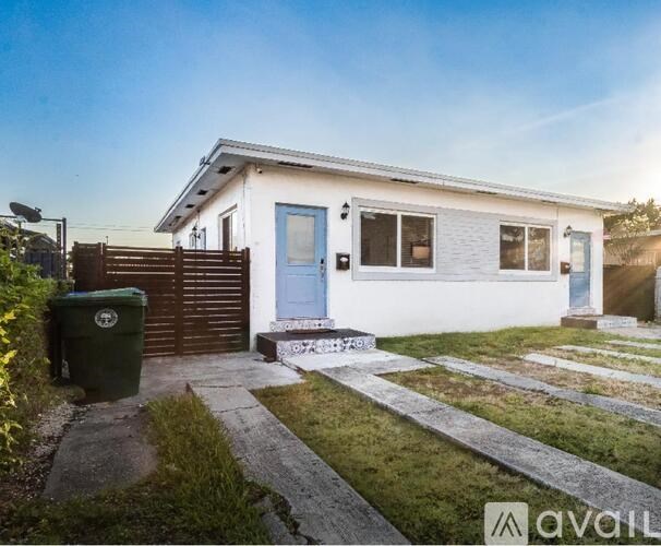 A small white house with a blue door and a green trash bin.