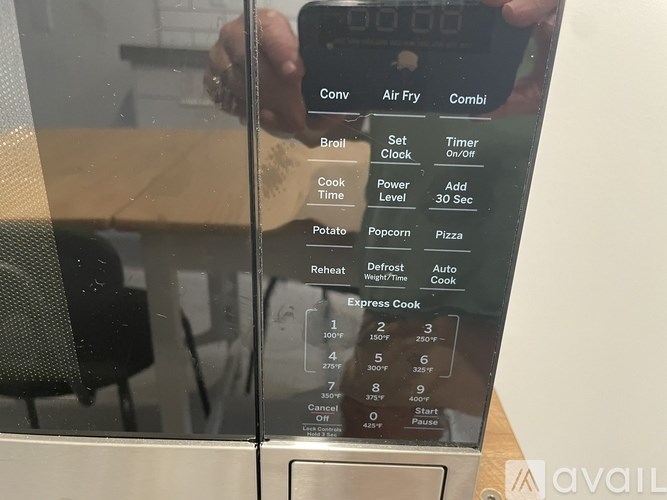 A person is holding a smartphone in front of a microwave with a digital display showing the time as 10:00.