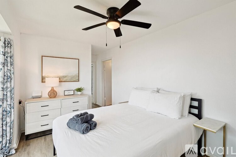 A bedroom with a white bed, a ceiling fan, and a nightstand with a lamp.