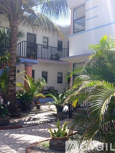 A white building with a balcony and a black railing is surrounded by palm trees.