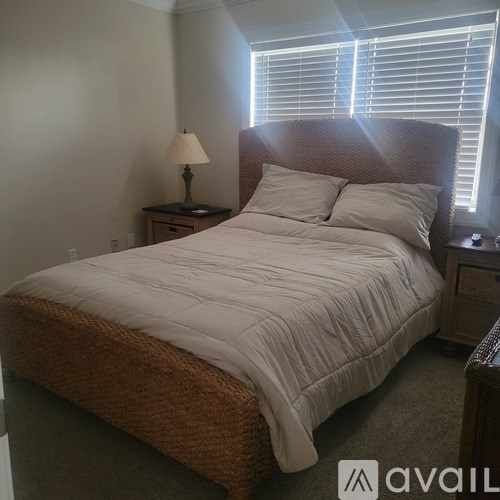 A bed with a white comforter and two pillows sits in a room with a lamp and window.
