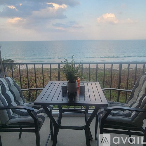 A table with two chairs and a potted plant on a balcony overlooking the ocean.