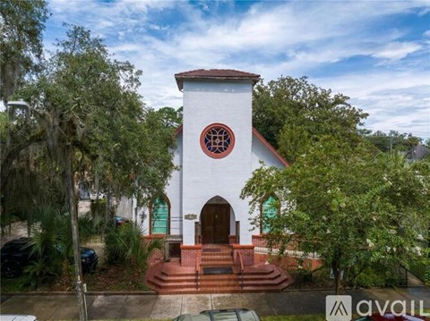 A white church with a red roof and a circular window is surrounded by trees.