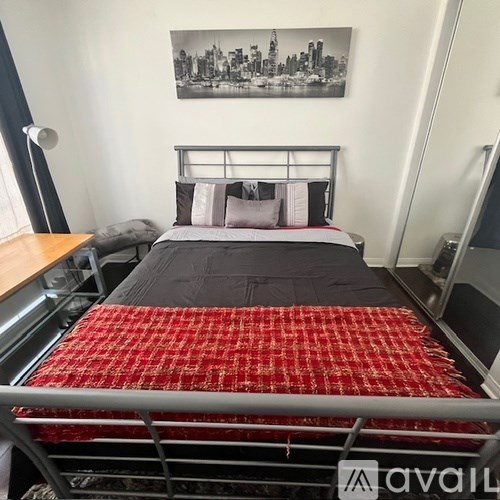 A bed with a red bedspread is in the foreground of a room with a cityscape picture on the wall.