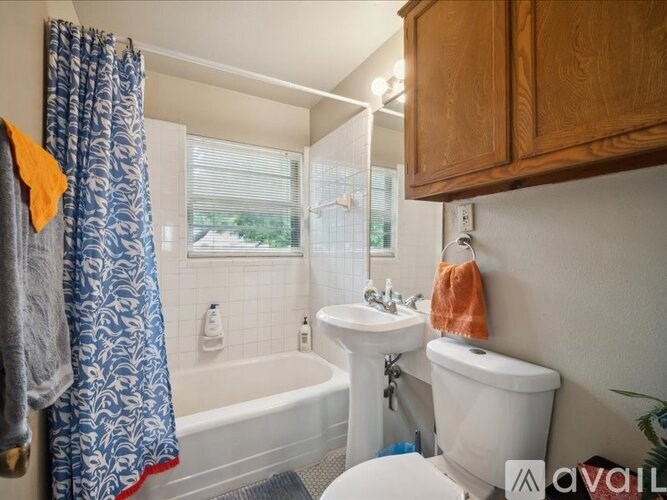 A bathroom with a white toilet, a white sink, a bathtub with a blue shower curtain, and a window with blinds.