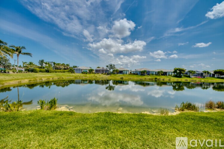 A serene landscape with a pond, grass, and buildings in the distance under a clear sky.