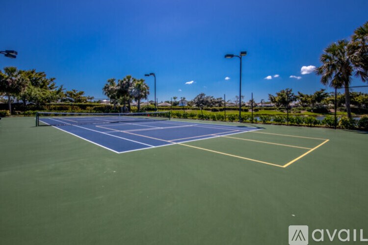 A tennis court with a blue top and white lines, surrounded by greenery and street lamps.