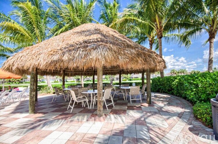 A thatched roof pavilion with chairs and tables is surrounded by palm trees.