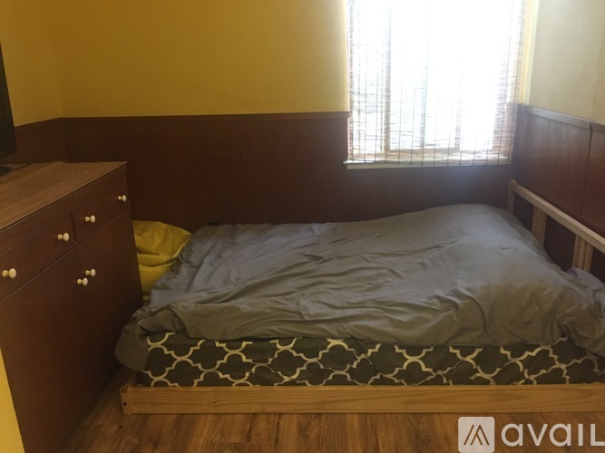 A bed with a grey cover and a green and white patterned pillow is in a room with wooden floors and a window with white curtains.