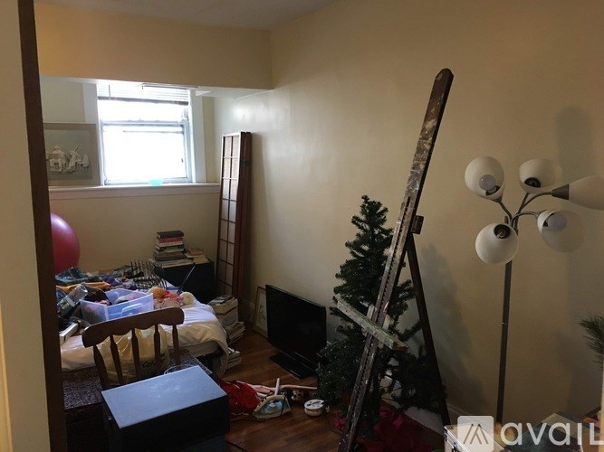 A room with a chair, a table, a bookshelf, a skateboard leaning against the wall, a small Christmas tree, and a floor lamp.