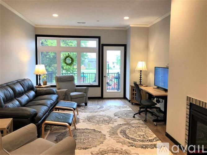 A living room with a black leather couch, a grey armchair, a wooden coffee table, a black office chair, a desk with a computer, a fireplace, and a large window with a view of a tree and a