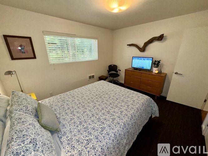 A bedroom with a bed, a lamp, a framed picture, a window with blinds, a wooden dresser with a laptop on top, and a decorative antler headboard.
