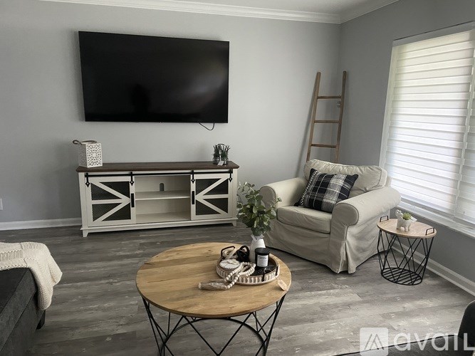 A living room with a flat screen TV mounted on the wall, a wooden coffee table in the center, a white armchair with a plaid pillow, and a wooden ladder leaning against the wall.