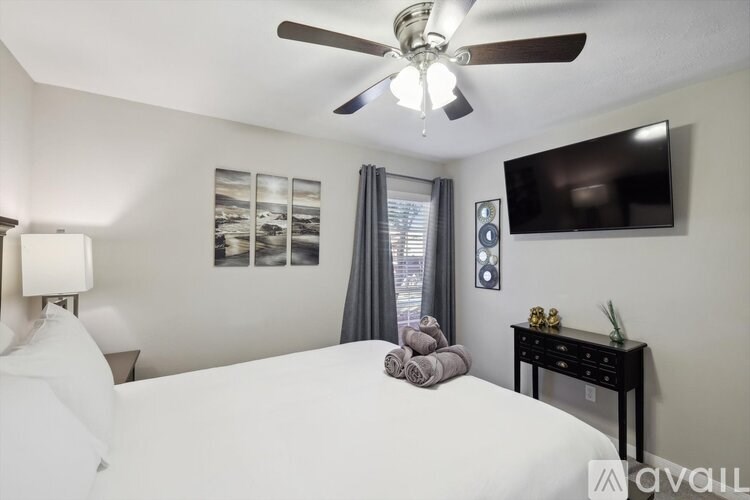 A bedroom with a bed, a ceiling fan, a nightstand, a table with a lamp, a window with curtains, and a wall-mounted TV.