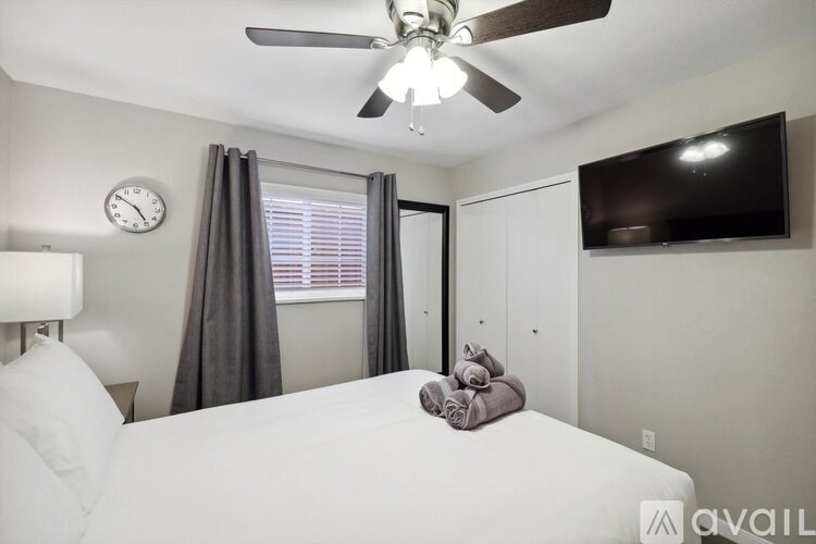 A bedroom with a bed, a ceiling fan, a clock, a window with curtains, a wall-mounted TV, and a wall-mounted shelf.