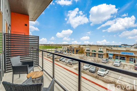 A balcony with chairs and a table overlooks a parking lot and buildings.
