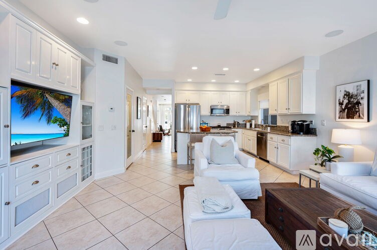 A modern kitchen with white cabinets and appliances, a TV displaying a beach scene, and a living area with a sofa and armchairs.