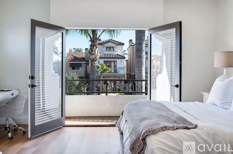 A bedroom with a large bed and a view of a balcony with a house and palm trees in the background.