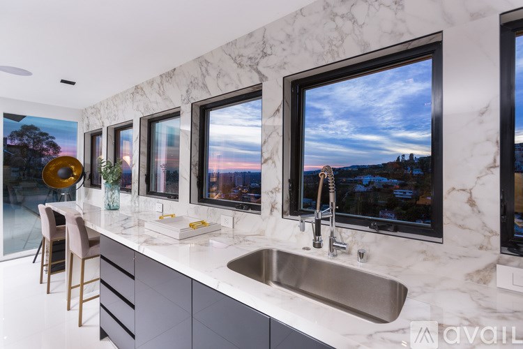A modern kitchen with marble countertops and a large window overlooking a sunset.