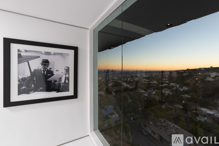 A framed black and white photo is hanging on a wall next to a window with a view of a cityscape at sunset.