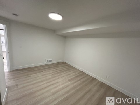 A room with wooden flooring and white walls, with a door on the left and a window on the right.