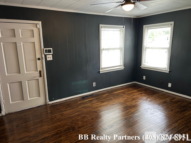 A room with a wooden floor, a ceiling fan, and two windows.