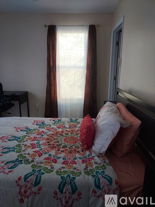 A bed with a floral patterned comforter and a red pillow is in the foreground of a room with a window and curtains.