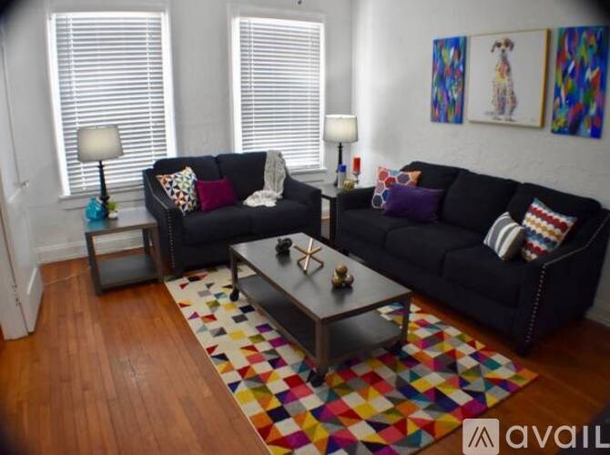 A black couch with colorful pillows sits in a room with wooden floors and white walls.